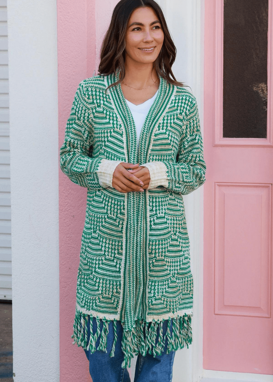 A woman wearing a long, open-front knit cardigan with green and white geometric patterns, featuring tassels at the hem. The boho cardigan is paired with a white top and high-waisted blue jeans.