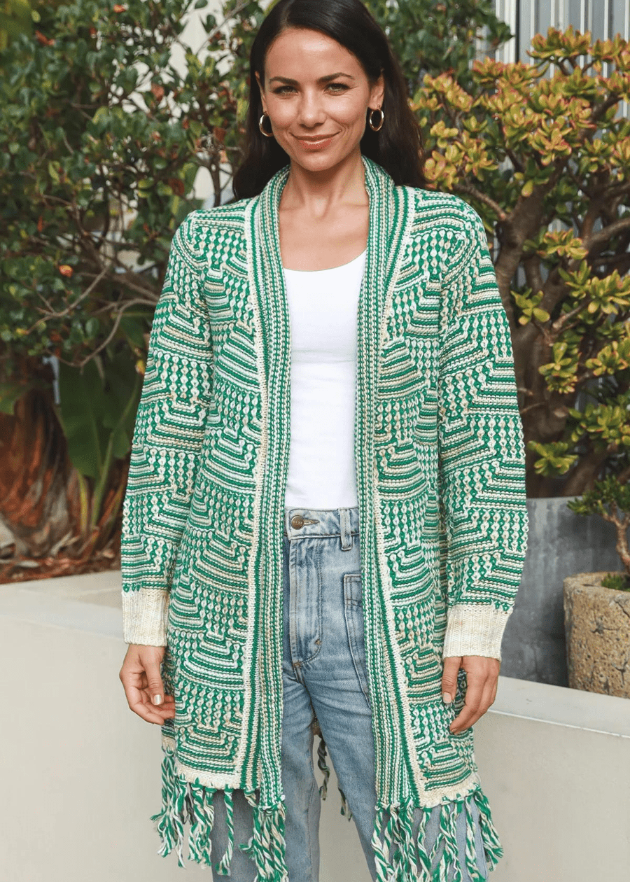 A woman wearing a long, open-front knit cardigan with green and white geometric patterns, featuring tassels at the hem. The boho cardigan is paired with a white top and high-waisted blue jeans.