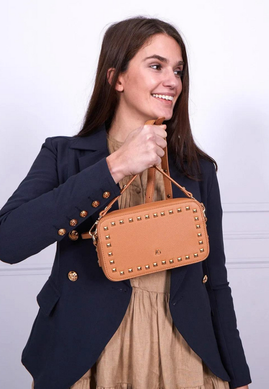 A woman holding a rectangular tan vegan leather handbag adorned with gold studs. The handbag exudes a chic and sophisticated vibe, perfect for elevating any outfit. With its stylish gold stud detailing, this vegan leather bag is a must-have accessory for those seeking a blend of elegance and modern edge. Adjustable straps to turn into a crossbody bag making it ideal for both casual and formal occasions