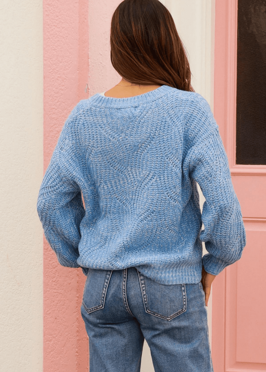 A woman wearing a light blue knit sweater with a subtle pattern on the back. The boho sweater is paired with high-waisted blue jeans. She is standing with her back to the camera, showing the detailed texture of the sweater. 