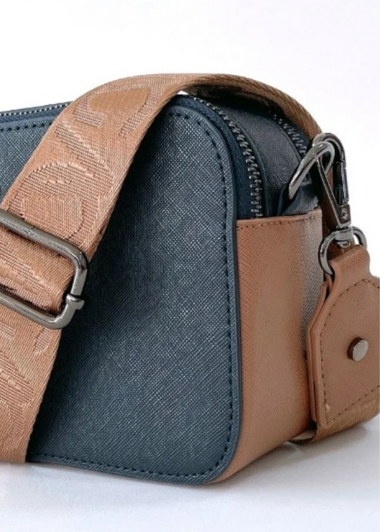 A dark blue vegan leather crossbody bag with silver zippers and a wide tan strap featuring a subtle embossed pattern. The bag has a small silver logo on the front and a minimalist design with a slim profile. The combination of navy blue and tan creates a sophisticated and versatile accessory.