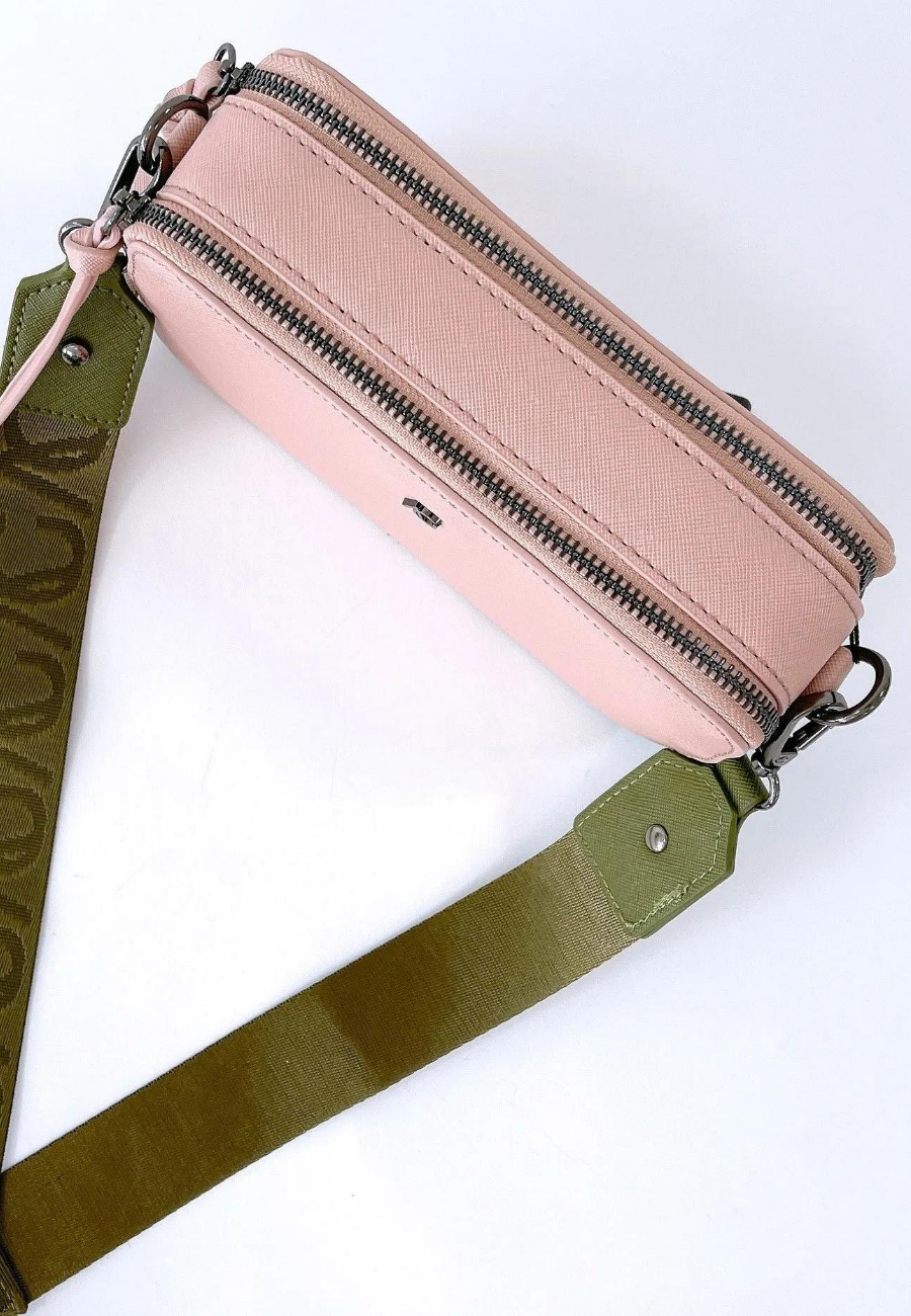 A pink vegan leather crossbody bag with silver zippers and a wide olive green strap featuring a subtle embossed pattern. The bag has a sleek, minimalist design with a small silver logo on the front. The combination of pastel pink and earthy green creates a stylish and versatile accessory.