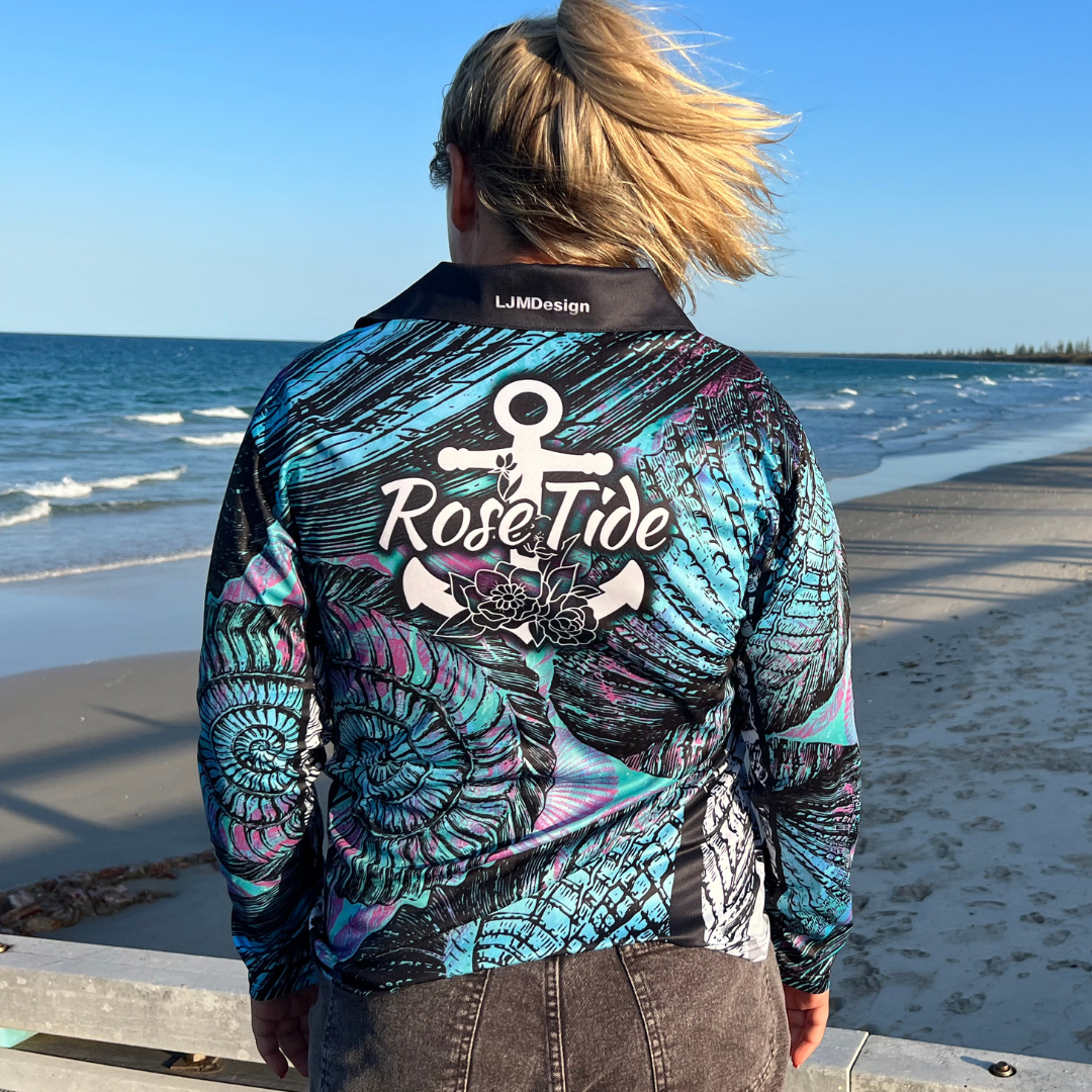 A woman wearing a A long sleeve women's fishing shirt featuring a detailed pattern of large, swirling seashells in shades of blue, teal, and purple with black accents. The "Rose Tide" logo with an anchor and floral design is on the chest. The shirt has a black collar and buttons, creating a striking and intricate oceanic design.