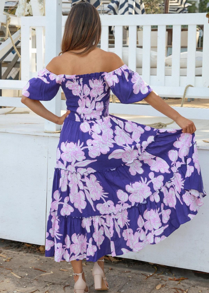 A woman wearing an off-the-shoulder Blue floral dress with pink and white flower prints. The dress features puff sleeves, a smocked bodice, and a tiered maxi skirt. The model is standing outdoors, smiling in a casual, relaxed setting.