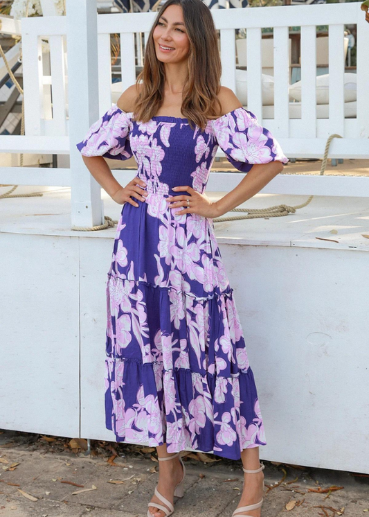 A woman wearing an off-the-shoulder Blue floral dress with pink and white flower prints. The dress features puff sleeves, a smocked bodice, and a tiered maxi skirt. The model is standing outdoors, smiling in a casual, relaxed setting.