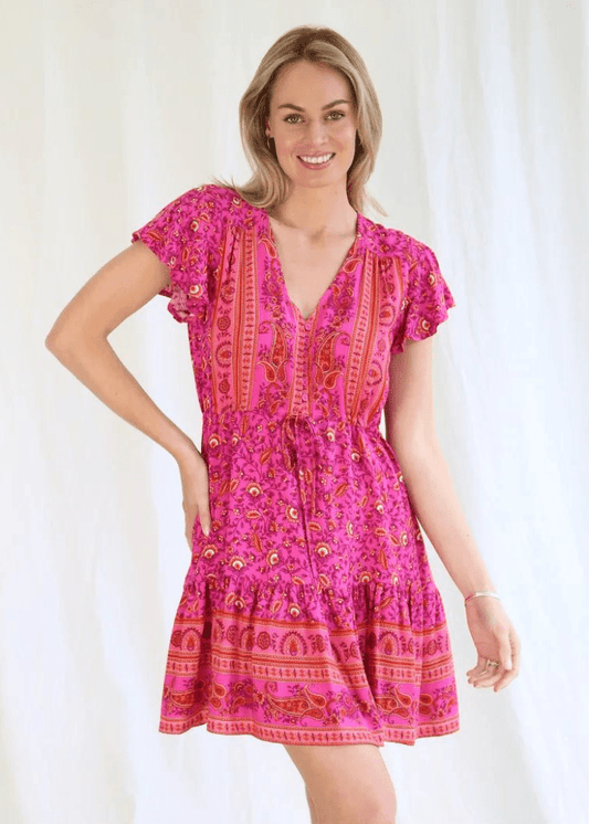 A woman wearing a mini dress with a rich, hot pink hue, embellished with ornate golden patterns that give it an exotic and luxurious feel. The dress has a deep V-neckline and short flutter sleeves, adding a playful yet elegant touch. The gathered waistline creates a flattering silhouette, and the ruffle at the hem enhances the dress's dynamic and festive vibe. It's a piece that stands out, perfect for those who love to make a bold fashion statement while maintaining a sense of femininity and fun.