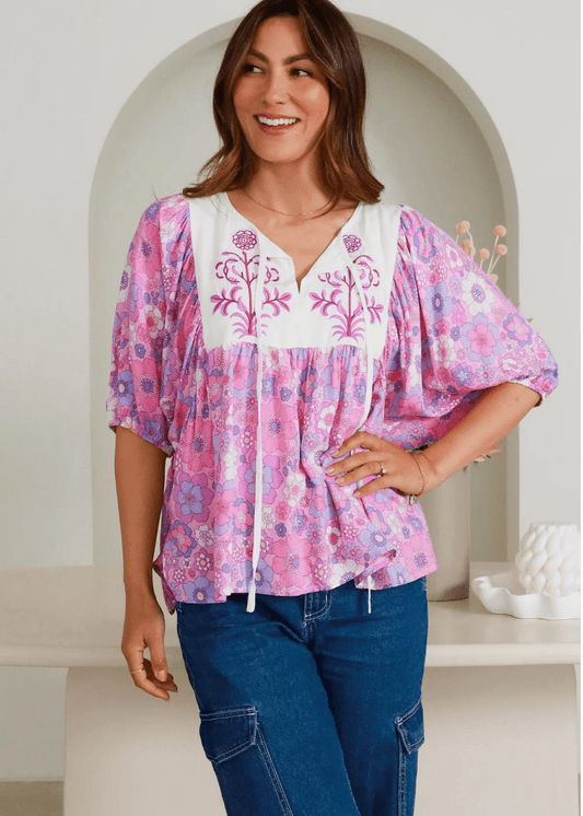 A pink and white floral embroidered top featuring a relaxed fit with flowing sleeves. The top has a V-neckline with intricate floral embroidery on the white yoke, complemented by a vibrant pink floral print throughout the rest of the fabric. 