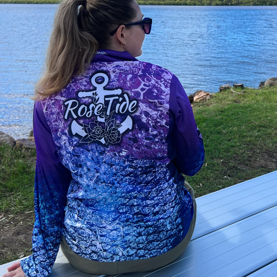 A woman wearing A long sleeve women's fishing shirt with a gradient design featuring shades of purple, blue, and pink, resembling ocean waves and fish scales. The "Rose Tide" logo with an anchor and floral design is on the chest. The left sleeve has the text "Follow the tide" written in white.