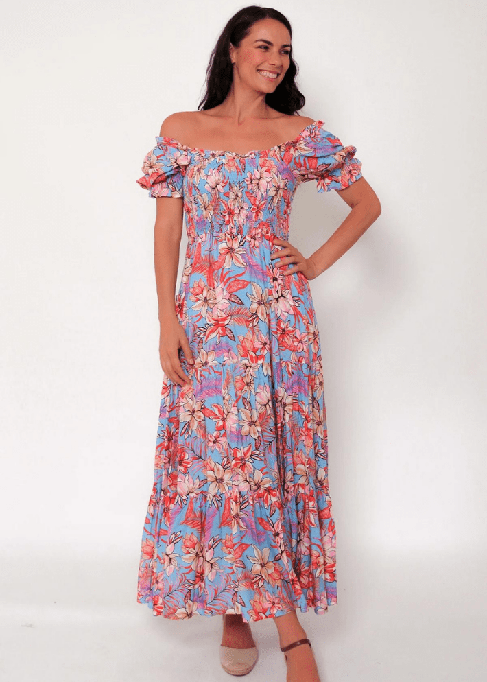 Women's off or on the shoulder maxi dress with flounce sleeves, combining a vivid floral print in pinks, oranges, and whites on a light blue base. It features a fitted bodice, a gently gathered waist, and a flowing tiered skirt. 