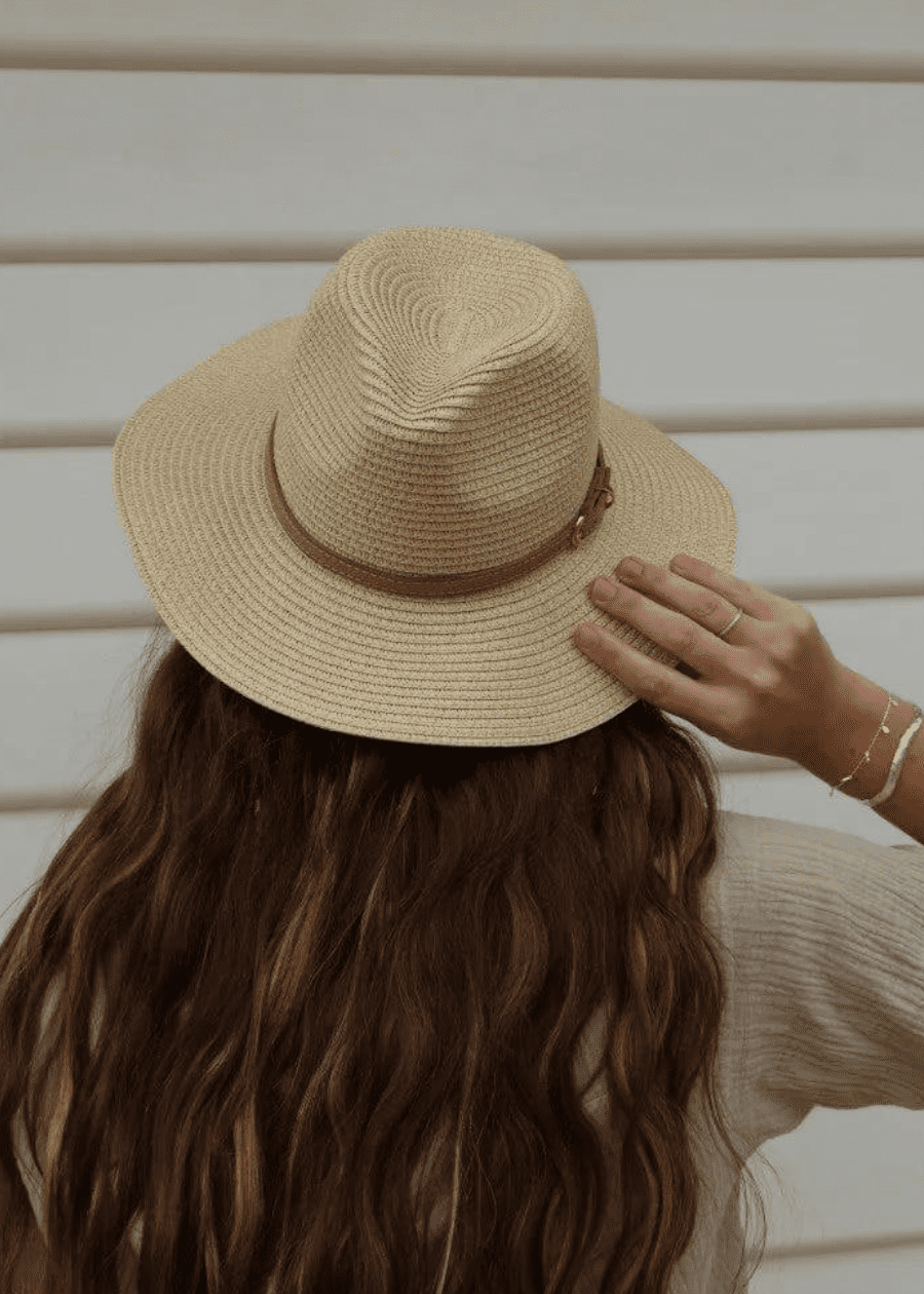 A woman with long, wavy hair seen from behind, accessories her outfit wearing a sand colour woven hat with a brown band and buckle, adding a casual, boho chic and stylish touch to her outfit.