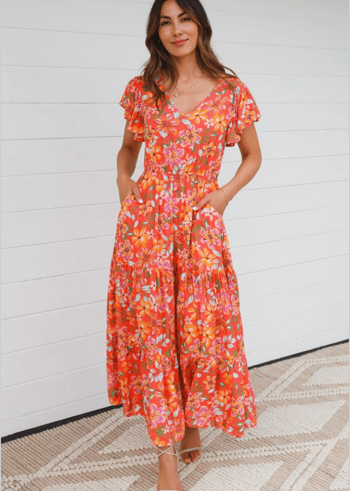 A woman wearing the Hazel maxi dress. The dress features an orange, yellow, purple, green, and white floral pattern on an orange background. The dress boasts flutter sleeves, a v-neckline, and a drawstring cinched waist leading to a tiered midi-length skirt that exudes summer vibes.