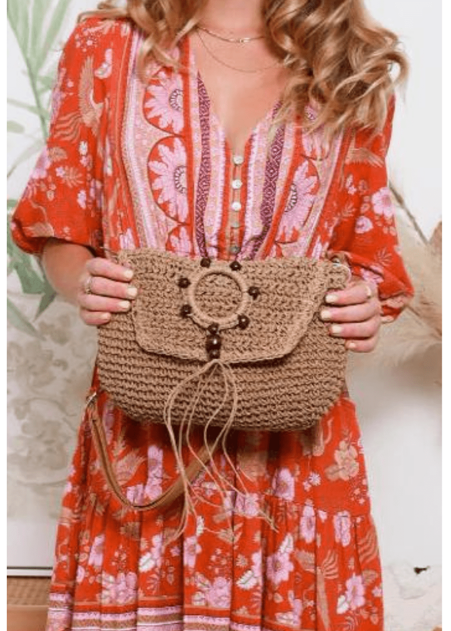 A woman holds a tan woven straw handbag with brown leather straps and wooden bead embellishments. She is smiling and standing outdoors, with greenery in the background. The handbag has a flap closure with a decorative circular design on the front.