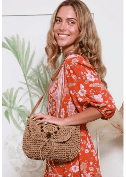 A woman holds a tan woven straw handbag with brown leather straps and wooden bead embellishments. She is smiling and standing outdoors, with greenery in the background. The handbag has a flap closure with a decorative circular design on the front.