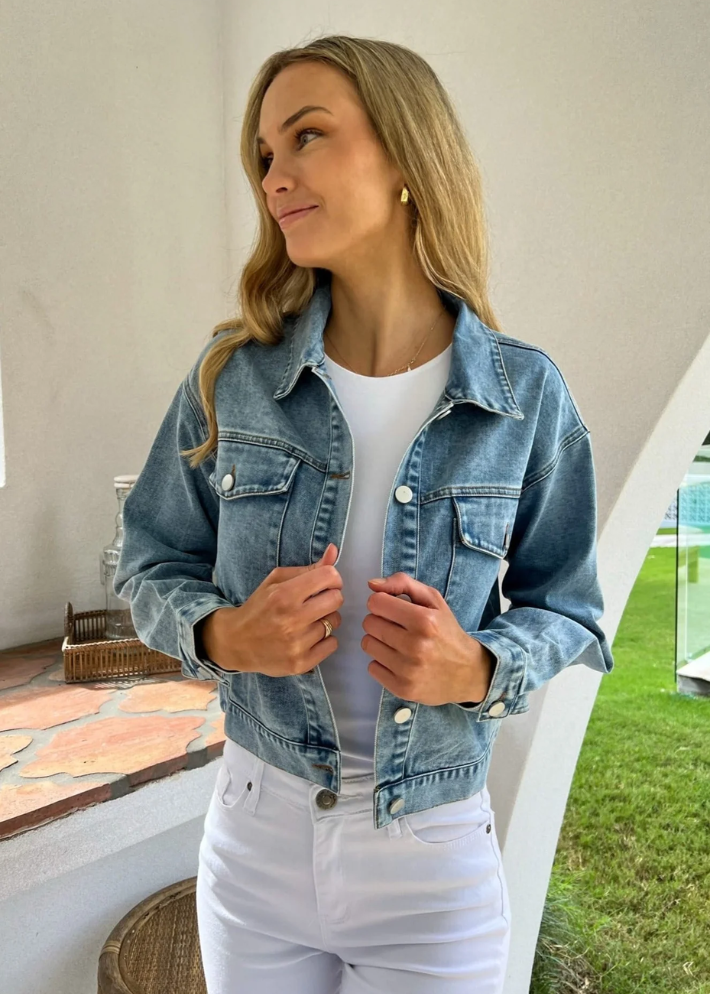 A close-up shot of a woman wearing a non-stretch, light blue denim jacket with pocket details over a white fitted top paired with white high-waisted jeans. The jacket has a relaxed fit and adjustable waistline buttons, creating a casual chic look.