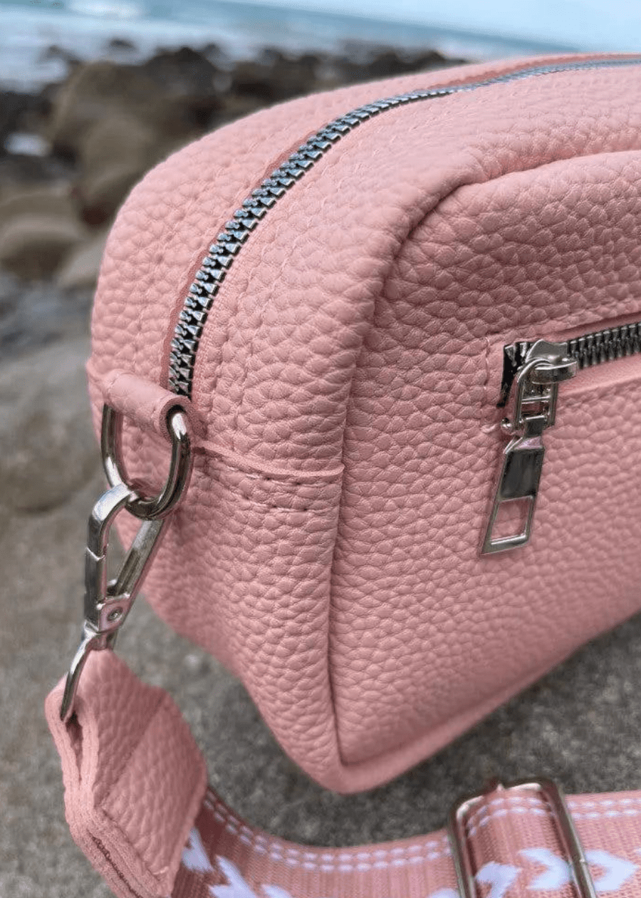 A pink crossbody bag made of vegan leather with a textured surface, featuring silver zippers and hardware. The bag is placed on a rocky surface with a blurred background, showcasing a pink and white, adjustable shoulder strap. 