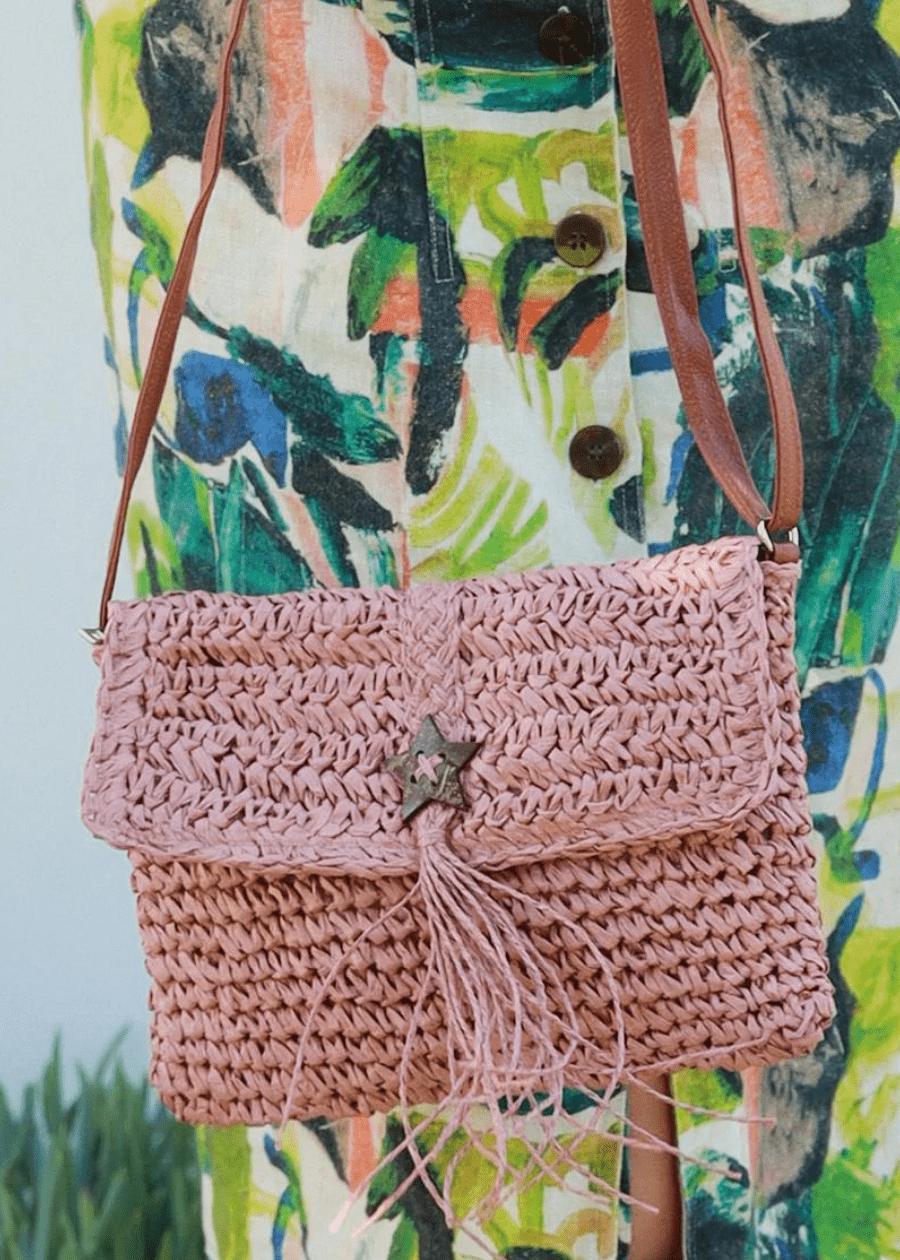 A close-up of a woman wearing a colourful, abstract-patterned dress holding a pink woven straw handbag with brown straps. The handbag features a flap closure with a star-shaped button and tassel detail, adding to its bohemian look. 