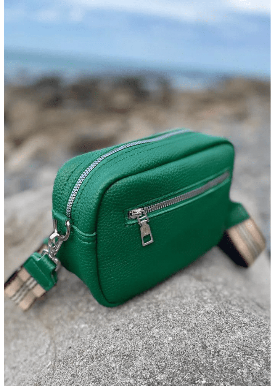 A green vegan leather crossbody bag with silver zippers and hardware, featuring a textured surface. The bag is placed on a rocky surface with a blurred coastal background, showcasing its adjustable, striped shoulder strap. The setting highlights the bag's vibrant colour and practical design, ideal for a stylish, casual look.