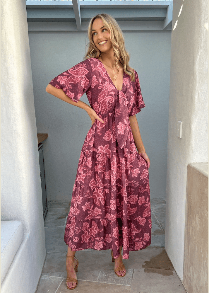 A woman wearing a Malbec Lily Maxi dress. The dress is a bohemian-style maroon dress adorned with a lighter pink floral pattern featuring flutter sleeves, a deep V-neckline, a tie front, and a gathered waistline that enhances the flowing skirt. 