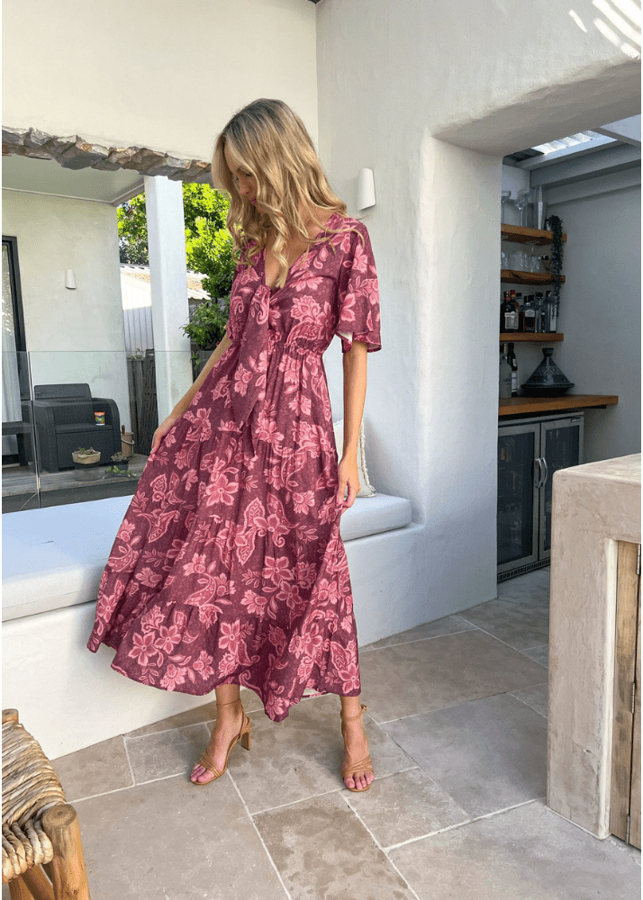 A woman wearing a Malbec Lily Maxi dress. The dress is a bohemian-style maroon dress adorned with a lighter pink floral pattern featuring flutter sleeves, a deep V-neckline, a tie front, and a gathered waistline that enhances the flowing skirt. 