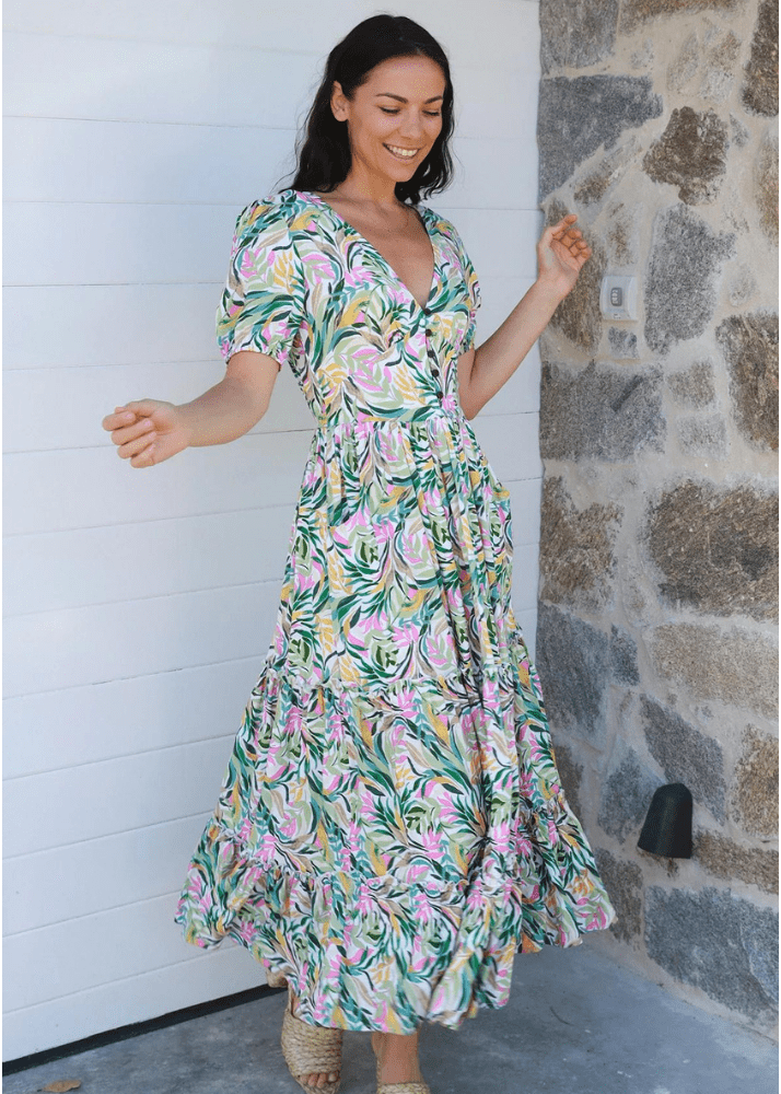 Woman in a full-length Ava & Mia tropical print maxi dress, sizes 8 to 16, perfect for summer. The dress features a blend of green, pink, and yellow foliage patterns on a lightweight rayon fabric, offering both comfort and style. It has a flattering V-neckline, short sleeves, 5 small buttons, cinched waist and a tiered skirt.