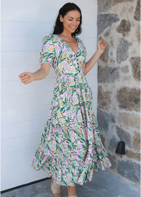 Woman in a full-length Ava & Mia tropical print maxi dress, sizes 8 to 16, perfect for summer. The dress features a blend of green, pink, and yellow foliage patterns on a lightweight rayon fabric, offering both comfort and style. It has a flattering V-neckline, short sleeves, 5 small buttons, cinched waist and a tiered skirt.