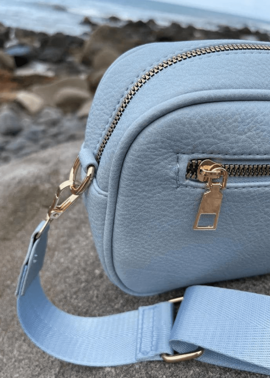 A light blue crossbody bag made of vegan leather with a textured surface, featuring gold zippers and hardware. The bag is placed on a rocky surface with a blurred background, showcasing a light blue, adjustable shoulder strap.