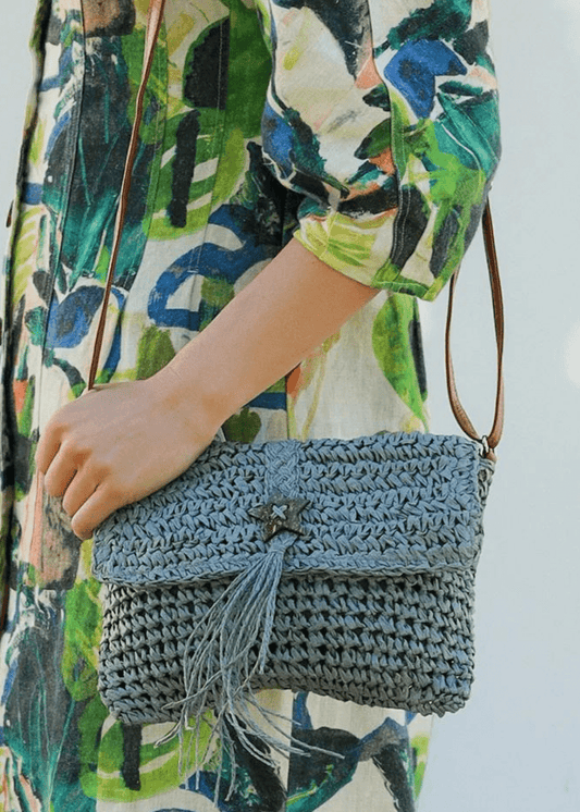 A close-up of a woman wearing a colorful, abstract-patterned dress holding a blue-grey woven straw handbag with brown straps. The handbag features a flap closure with a star-shaped button and tassel detail, adding to its bohemian look