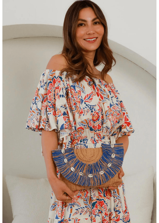 A woman holds a tan woven clutch adorned with blue fringe and white shell accents. The clutch has a semi-circular design and is paired with a white dress featuring a vibrant, multicolored floral and leaf pattern. The accessory and dress together create a bohemian, beachy look.