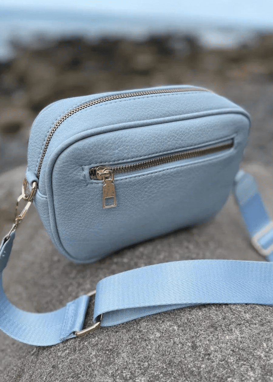 A light blue crossbody bag made of vegan leather with a textured surface, featuring gold zippers and hardware. The bag is placed on a rocky surface with a blurred background, showcasing a light blue, adjustable shoulder strap. 