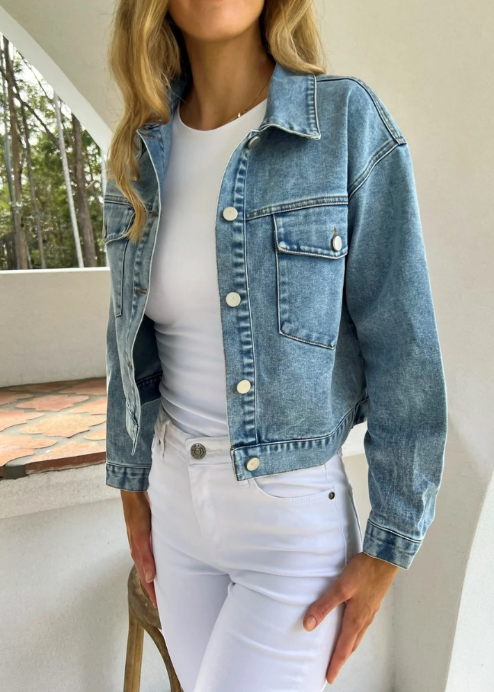 A close-up shot of a woman wearing a non-stretch, light blue denim jacket with pocket details over a white fitted top paired with white high-waisted jeans. The jacket has a relaxed fit and adjustable waistline buttons, creating a casual chic look.