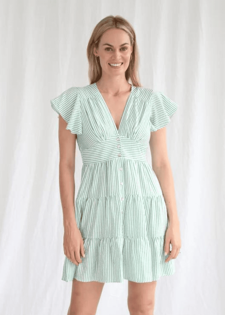 A woman wearing a light green and white striped dress with a flattering v-neckline and button details that run down the front, adding a touch of classic charm. The flutter sleeves and gathered waist leading to a flared skirt provide a soft, feminine silhouette. This dress would be perfect for a casual spring or summer day, suitable for a variety of occasions where comfort and a light-hearted aesthetic are desired.