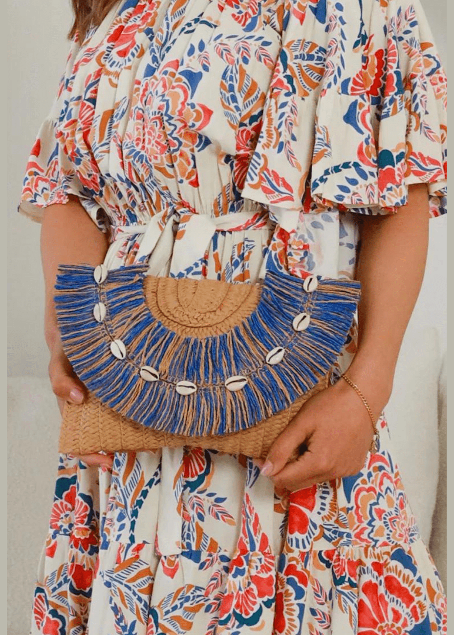 A woman holds a tan woven clutch adorned with blue fringe and white shell accents. The clutch has a semi-circular design and is paired with a white dress featuring a vibrant, multicolored floral and leaf pattern. The accessory and dress together create a bohemian, beachy look.