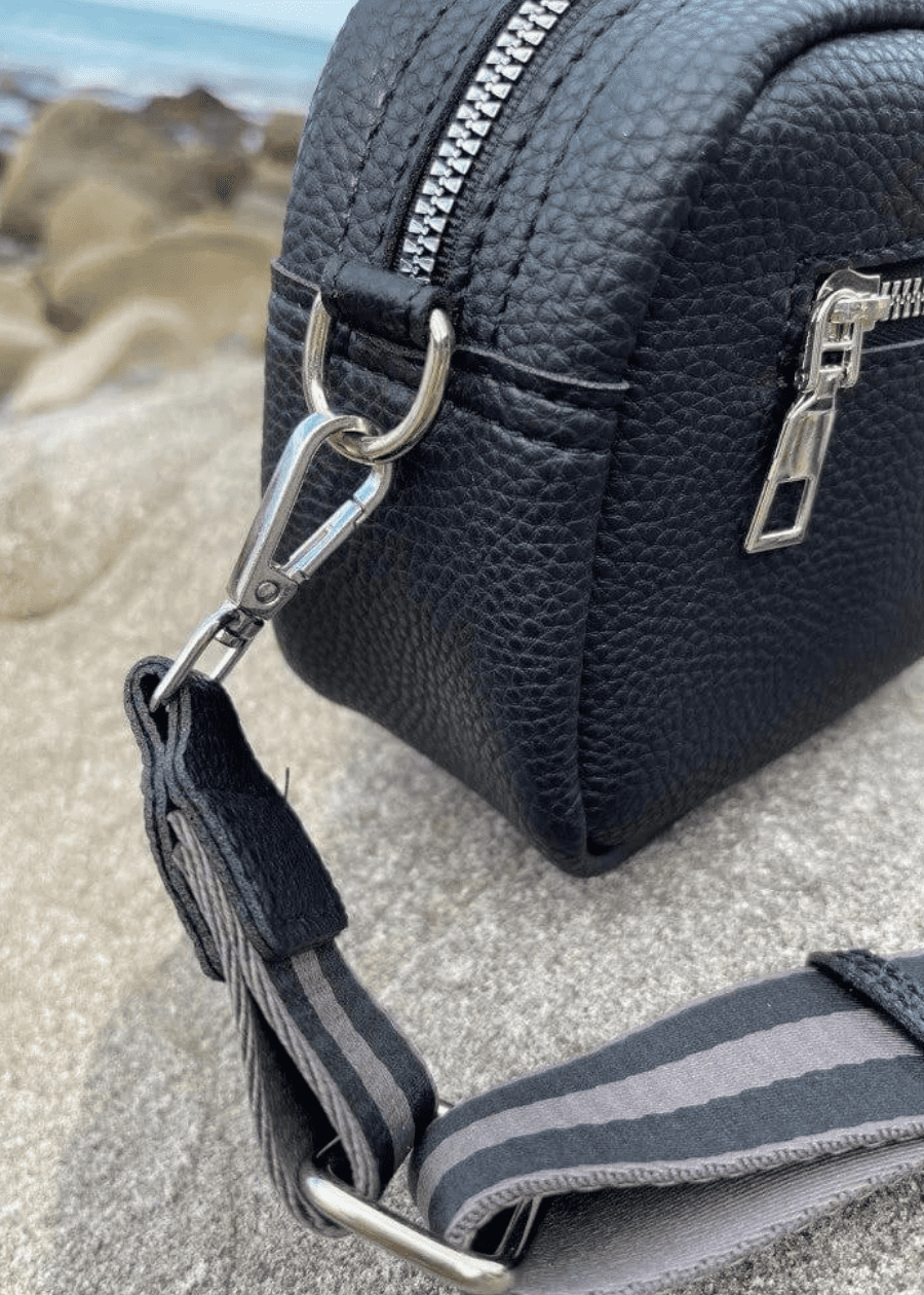 A black crossbody bag made of vegan leather with a textured surface, featuring silver zippers and hardware. The bag is placed on a rocky surface with a blurred background, showcasing a striped, adjustable shoulder strap. 