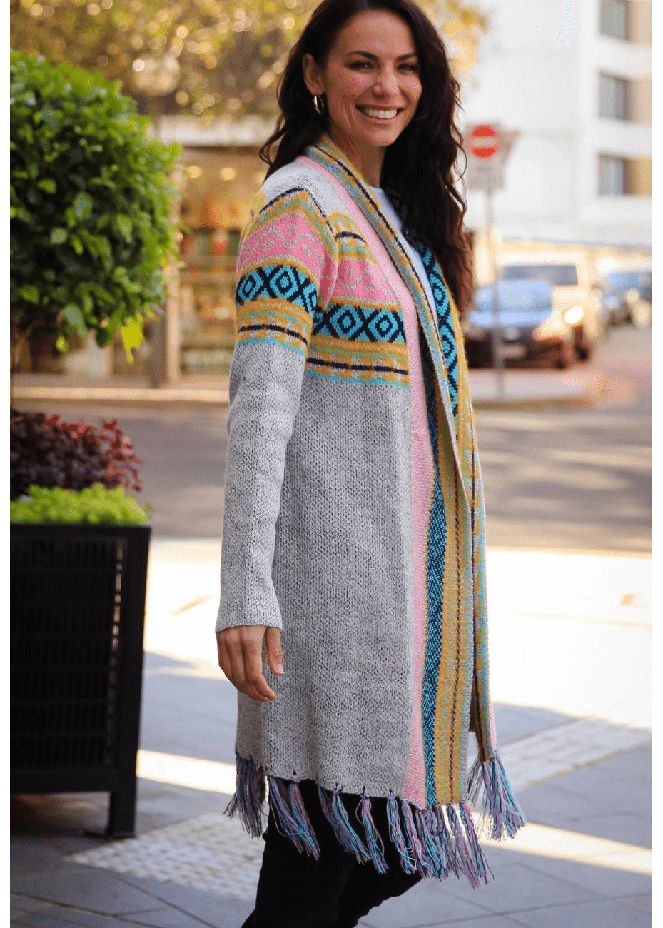 A woman smiles while wearing a long, patterned cardigan with grey, blue, pink, and yellow stripes, featuring diamond motifs on the shoulders and fringes at the hem.