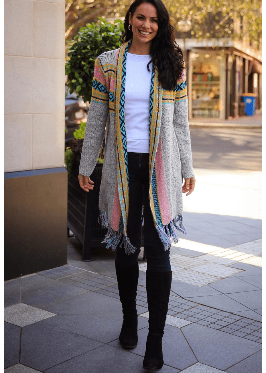 A woman smiles while wearing a long, patterned cardigan with grey, blue, pink, and yellow stripes, featuring diamond motifs on the shoulders and fringes at the hem.