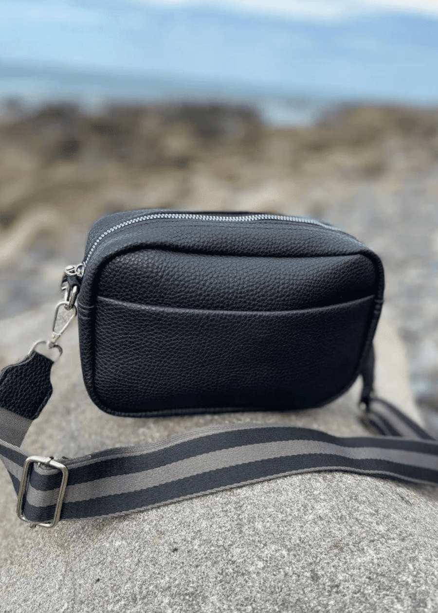 A black crossbody bag made of vegan leather with a textured surface, featuring silver zippers and hardware. The bag is placed on a rocky surface with a blurred background, showcasing a striped, adjustable shoulder strap. 