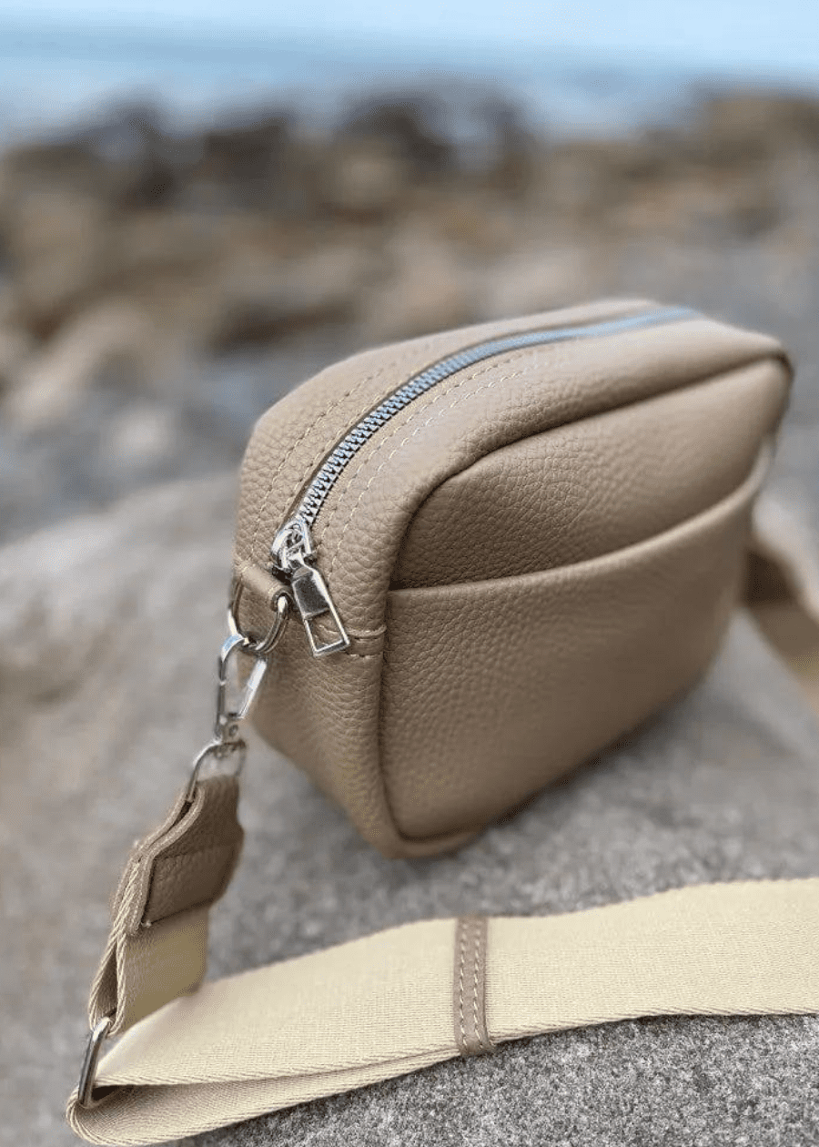 A beige crossbody bag made of vegan leather with a textured surface, featuring silver zippers and hardware. The bag is placed on a rocky surface with a blurred background, showcasing a adjustable shoulder strap. 