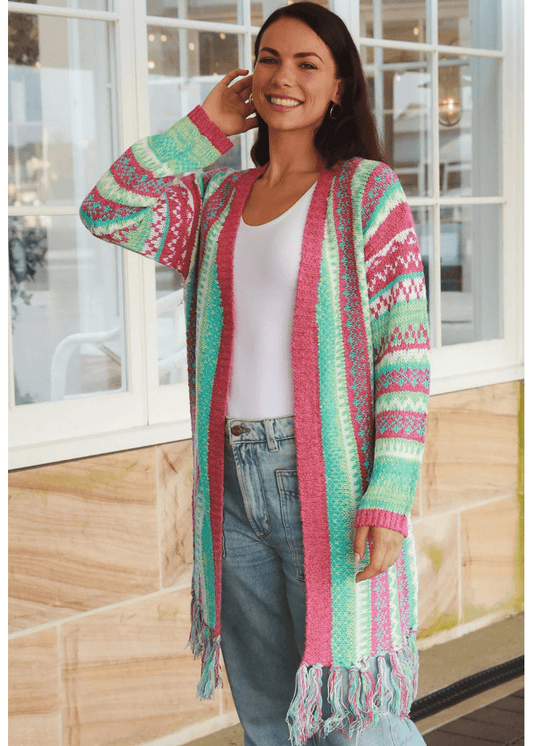 A woman wearing a long, open-front knit cardigan with colourful, geometric patterns in shades of green, pink, white, and blue. The boho cardigan has fringes at the hem and is paired with a white top and high-waisted blue jeans. 