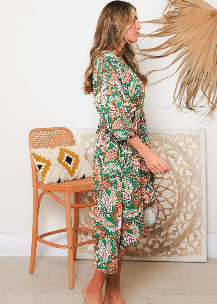 Women's Knee-length wrap dress featuring a tropical leaf print in shades of green, orange, purple and white on a back ground of greenwith a V-neckline, material waist tie and three-quarter sleeves 