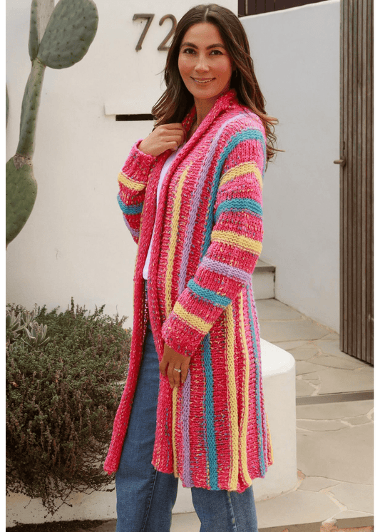 A woman wearing a long, open-front cardigan with vibrant, multicolored stripes in shades of pink, yellow, blue, and purple. She pairs the cardigan with a white top and blue jeans. The bohemian-inspired cardigan adds a playful and cozy touch to her casual outfit, creating a cheerful and stylish look.