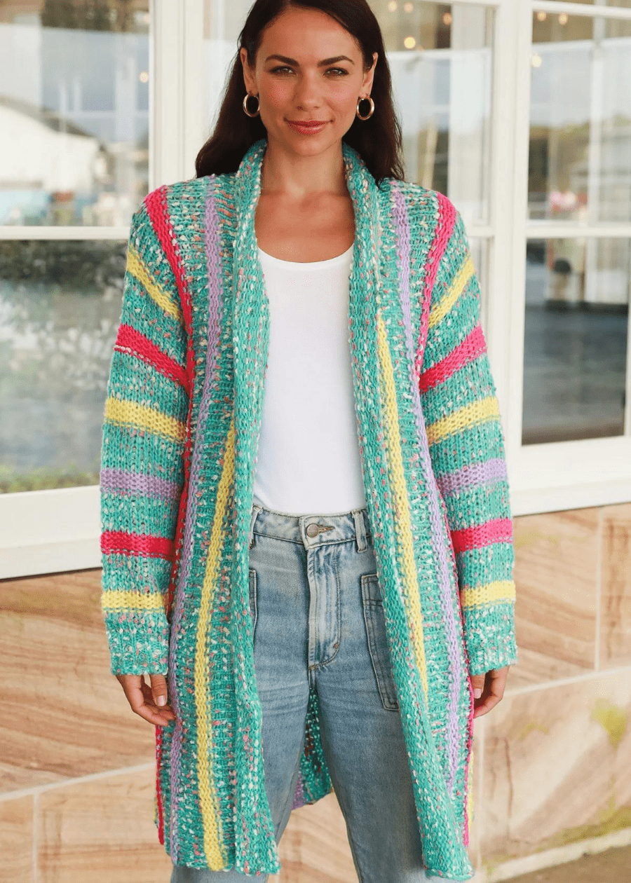 A woman wearing a long, knit open-front cardigan with colorful, striped patterns in shades of green, yellow, pink, and purple. She paired the boho cardigan with a white top and high-waisted blue jeans. 