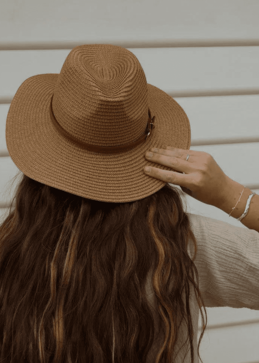 A woman with long, wavy hair seen from behind, accessories her outfit wearing a tan woven hat with a brown band and buckle, adding a casual, boho chic and stylish touch to her outfit. 