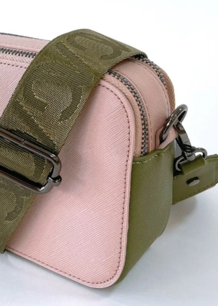 A pink vegan leather crossbody bag with silver zippers and a wide olive green strap featuring a subtle embossed pattern. The bag has a sleek, minimalist design with a small silver logo on the front. The combination of pastel pink and earthy green creates a stylish and versatile accessory.