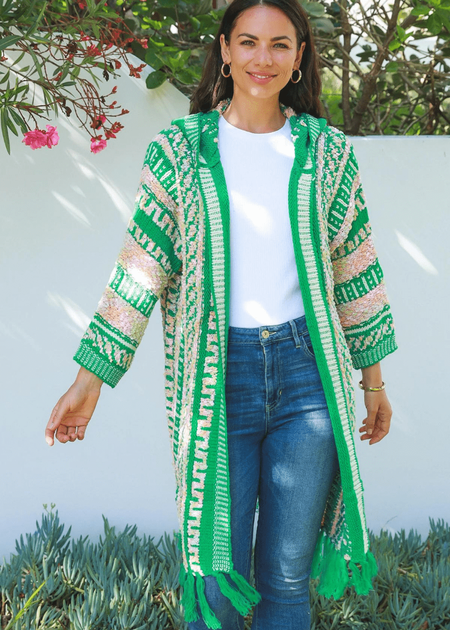 A woman wearing a long, open-front knit cardigan with green, white, and peach patterns, featuring tassels at the hem. The boho cardigan is paired with a white top and high-waisted blue jeans. She is smiling and standing outdoors near a white wall with greenery and flowers in the background, creating a fresh and vibrant look.