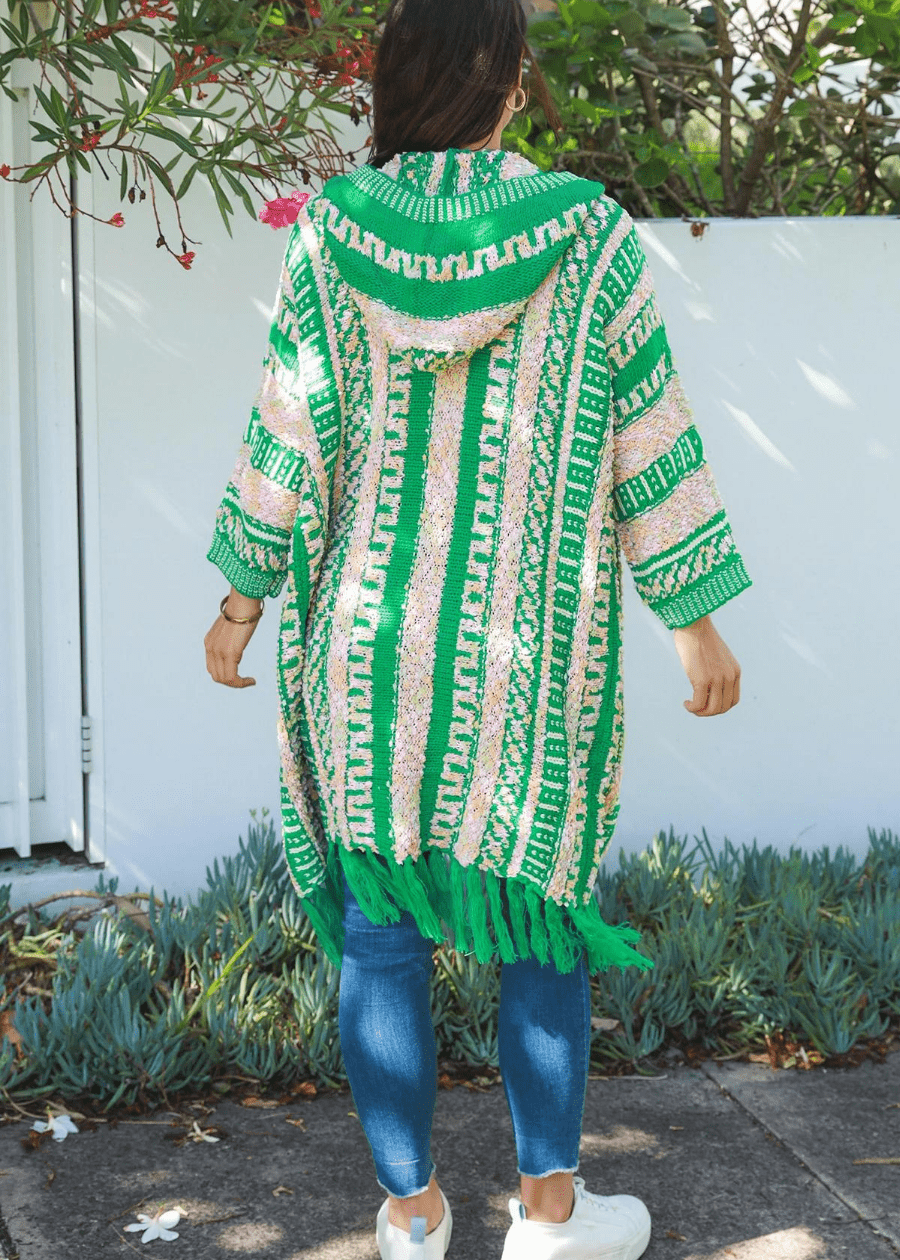 A woman wearing a long, open-front cardigan with green, white, and peach patterns, featuring tassels at the hem and a hood. The boho cardigan is paired with a white top and high-waisted blue jeans. 