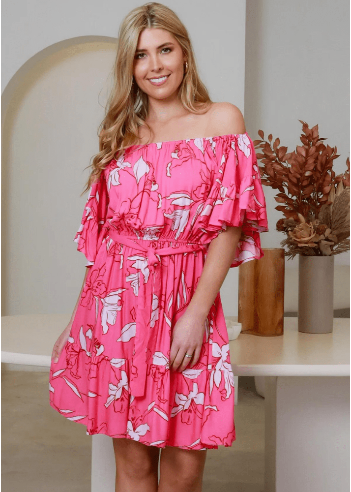 A woman wearing an off-the-shoulder, short, ruffled dress with a cinched waist in a vibrant pink hue adorned with large white floral patterns. The playful design features flowing ruffles along the neckline and hem, creating a breezy, summery look.  The Carly Mini Dress is from Ava & Mia  available in sizes 8-14