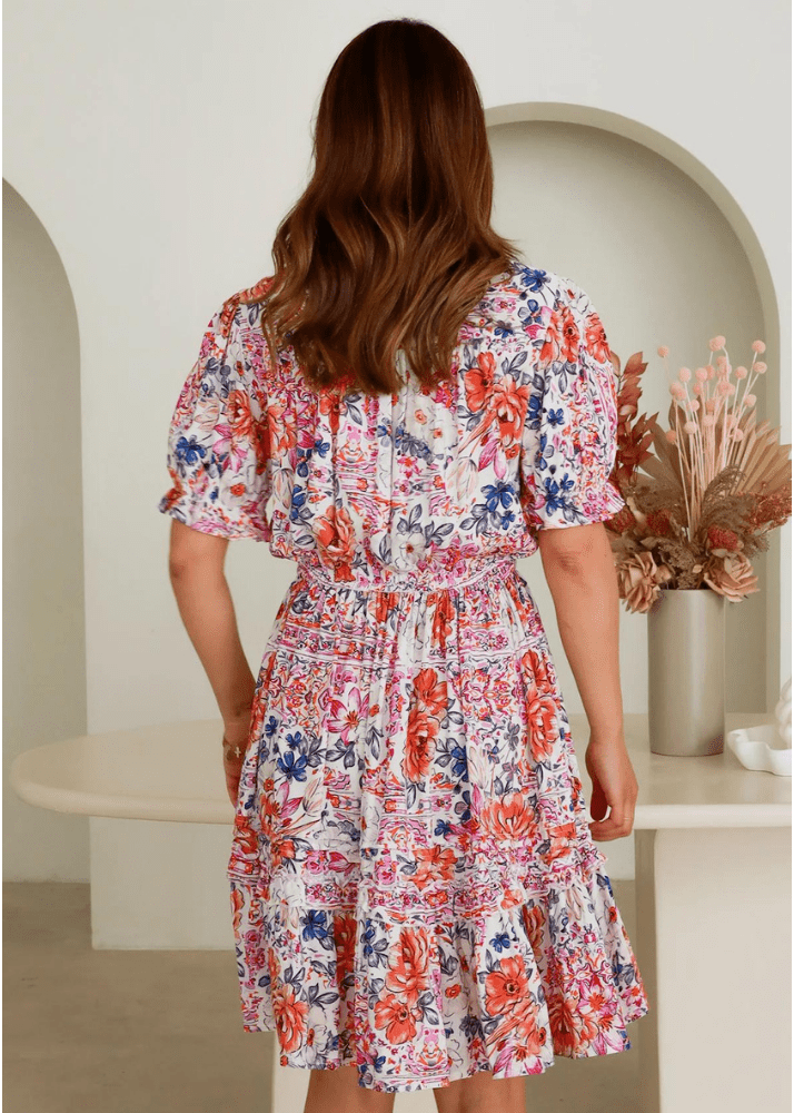 The dress features beautiful coral, blush, and sapphire florals on a soft white background. Each bloom is crafted to capture the wonder of a blooming garden. It's flirty and fun, with playful ruffles at the hem that add a whimsical touch to your outfit. It also has a perfect fit – cinched at the waist with a comfortable tie, creating a silhouette