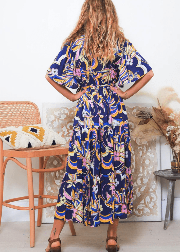 A woman dons a bohemian summer navy blue midi dress adorned with a lively pattern of yellow, pink, and white florals. The dress is designed with short sleeves, a V-neckline, and a waist tie that elegantly cinches at the waist, enhancing the figure. The skirt part of the dress flows gently to mid-calf length, adding to the garment's breezy, summery charm. 