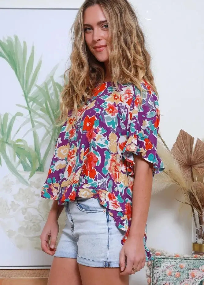 Loose-fitting blouse with a bold floral print in vivid colors of red, orange, purple, and turquoise. The blouse has a round neckline, elbow-length sleeves with a slight ruffle, and a peplum hem