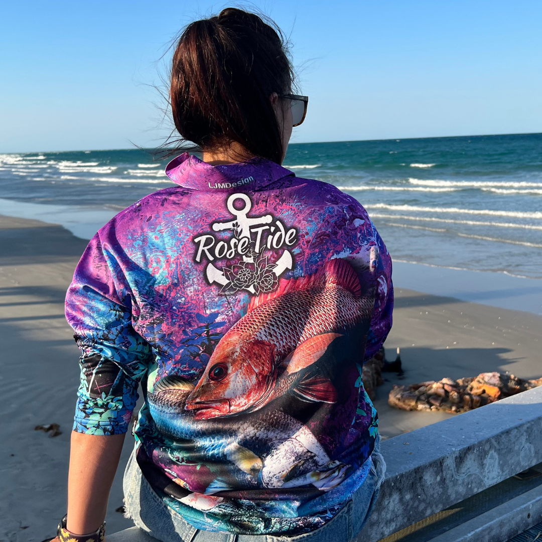 a woman wearing A long sleeve fishing shirt with a vibrant, abstract background in purple, blue, and pink tones. The design features a barra, mangrove jack and a crocodile prominently on the front, along with the "Rose Tide" logo on the chest, which includes an anchor and rose